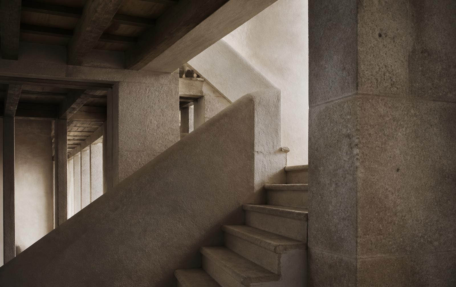 An interior stone stairwell with repeating beams and raking daylight, the structure of the building plainly visible.